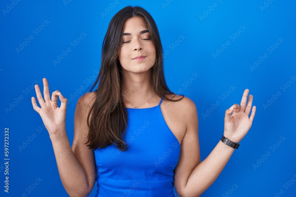 Fototapeta premium Hispanic woman standing over blue background relaxed and smiling with eyes closed doing meditation gesture with fingers. yoga concept.