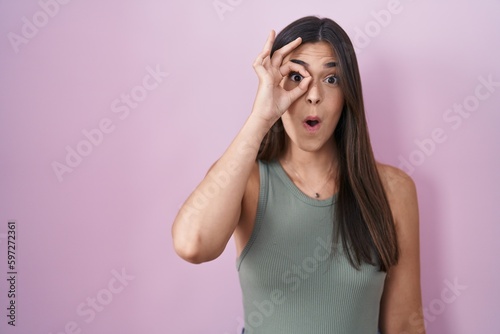 Hispanic woman standing over pink background doing ok gesture shocked with surprised face, eye looking through fingers. unbelieving expression.