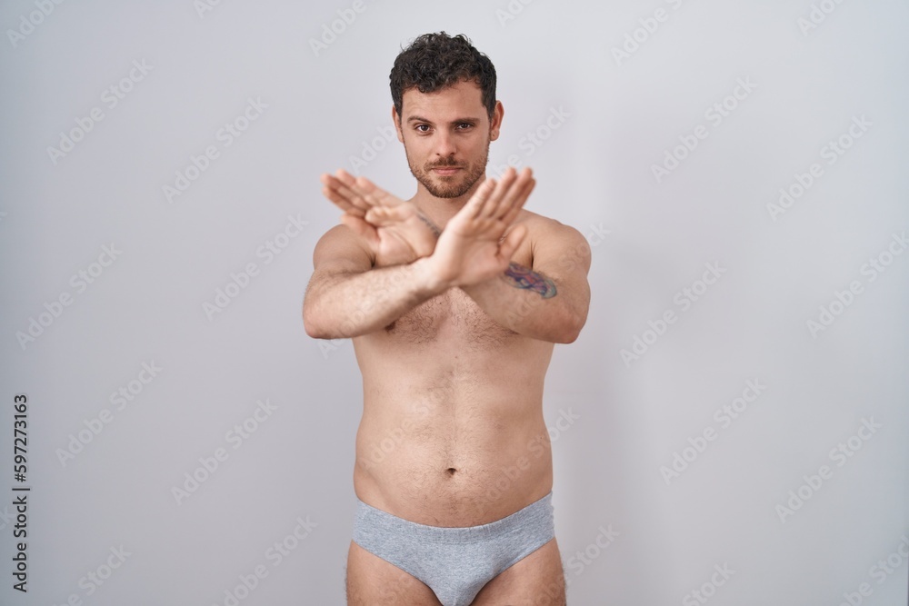Young hispanic man standing shirtless wearing underware rejection expression crossing arms and palms doing negative sign, angry face