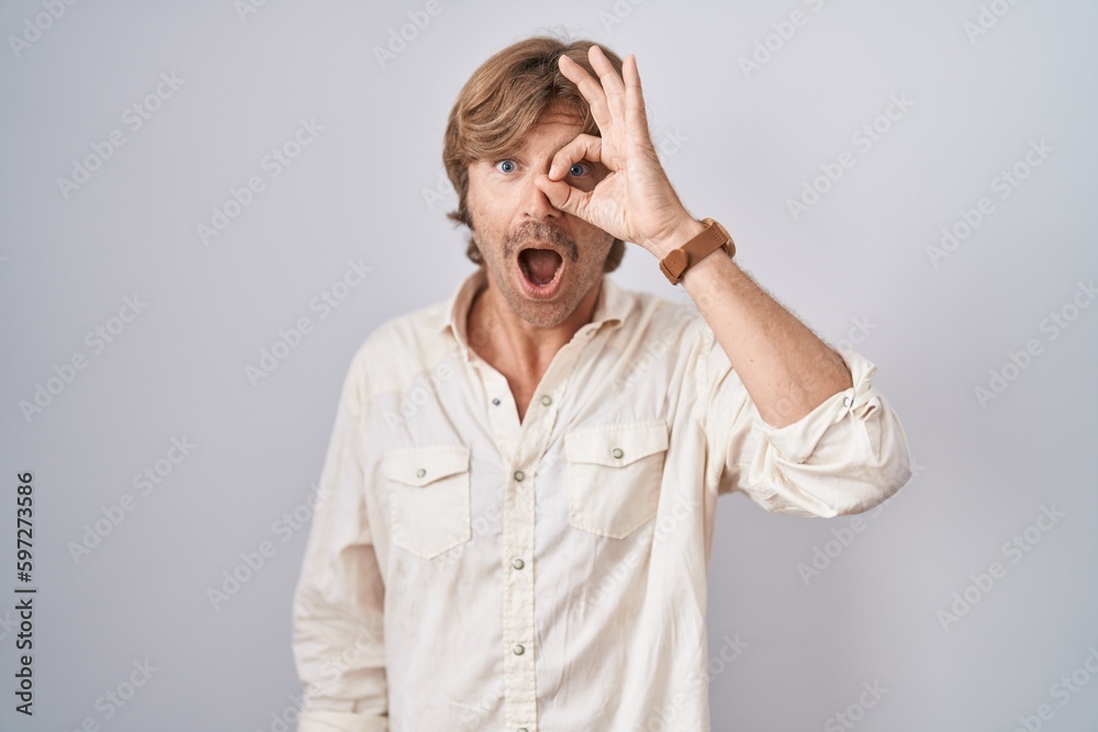 Middle age man standing over isolated background doing ok gesture shocked with surprised face, eye looking through fingers. unbelieving expression. Middle age man standing over isolated background doing ok gesture shocked with surprised face, eye looking through fingers. unbelieving expression.