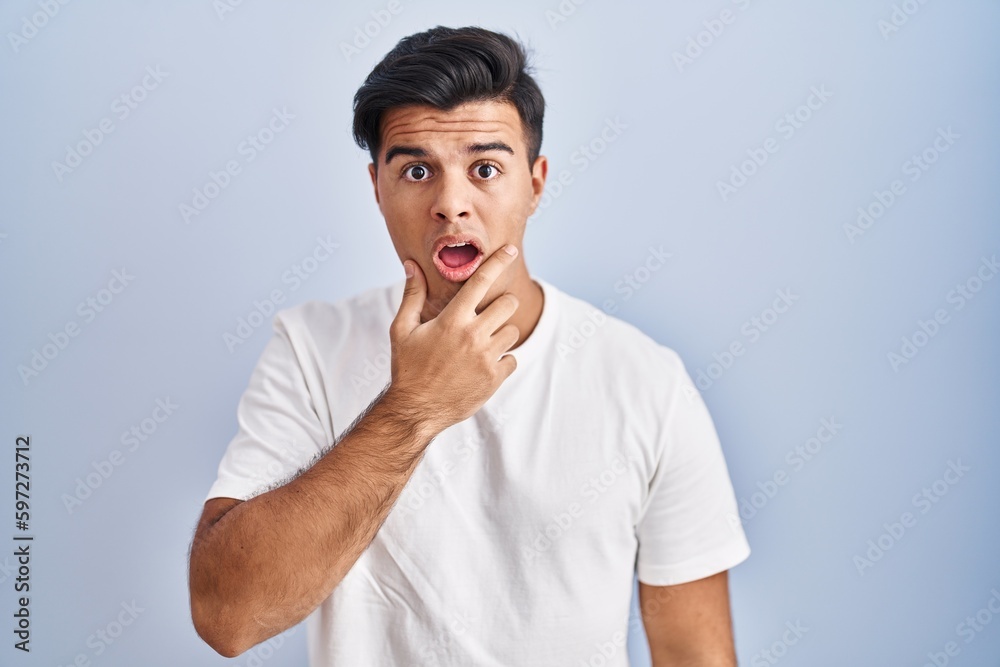 Hispanic man standing over blue background looking fascinated with disbelief, surprise and amazed expression with hands on chin Hispanic man standing over blue background looking fascinated with disbelief, surprise and amazed expression with hands on chin