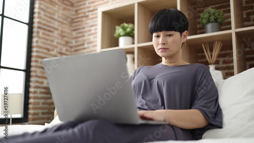 Young chinese woman using laptop sitting on bed at bedroom