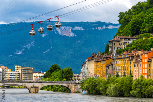Isere river in Grenoble city skyline, Auvergne-Rhone-Alpes region, France. Pont Marius-Gontard bridge, Grenoble-Bastille Cable car (Telepherique) and Alps mountains on background © katatonia
