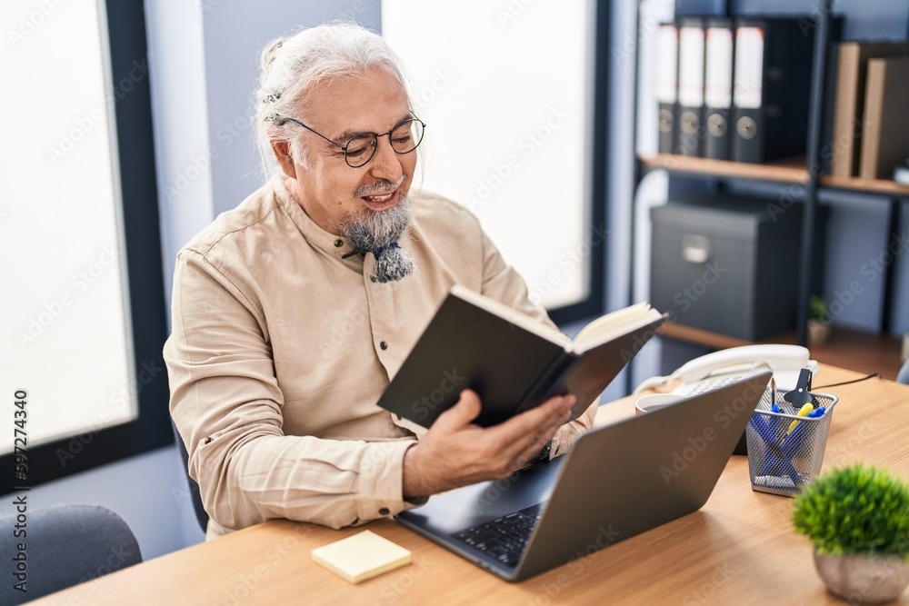 Middle age grey-haired man business worker using laptop reading book at office Middle age grey-haired man business worker using laptop reading book at office