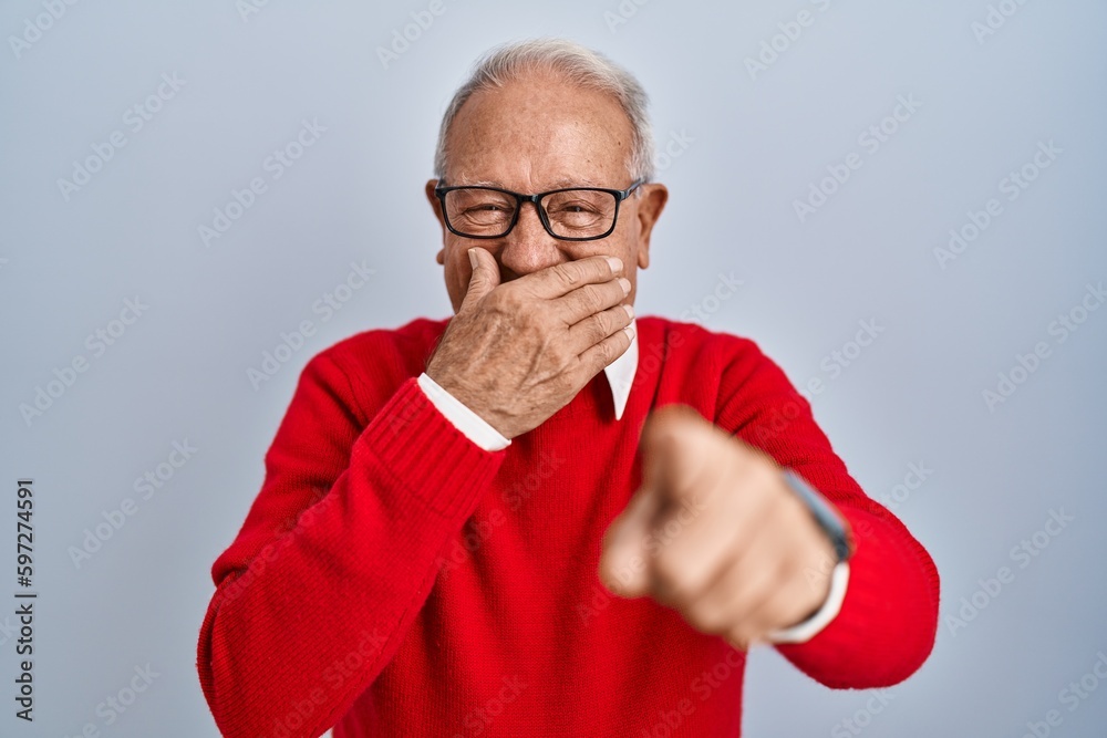 Senior man with grey hair standing over isolated background laughing at you, pointing finger to the camera with hand over mouth, shame expression Senior man with grey hair standing over isolated background laughing at you, pointing finger to the camera with hand over mouth, shame expression