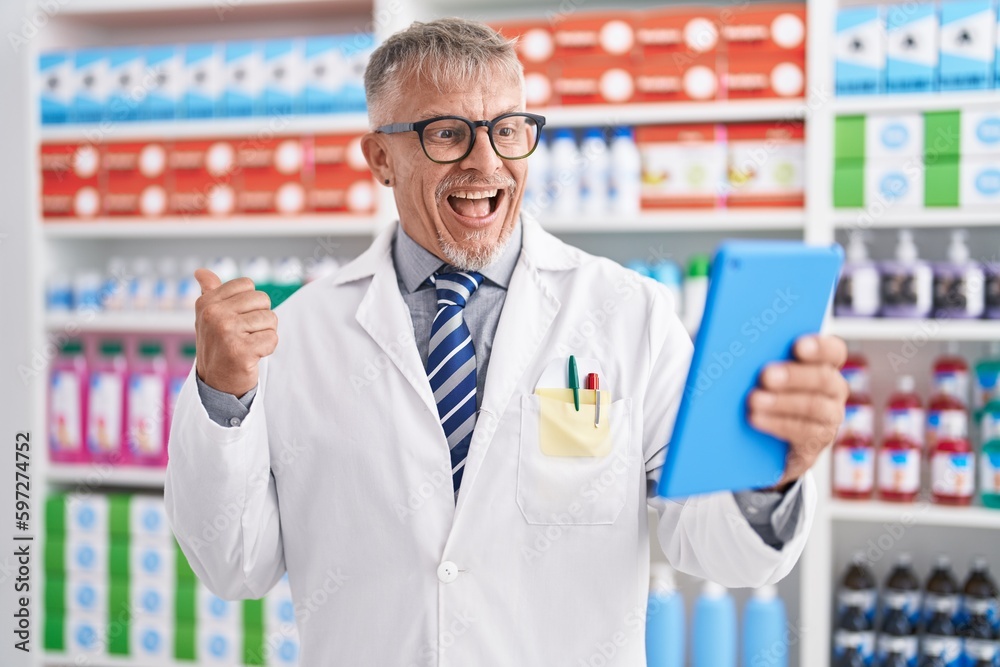 Hispanic man with grey hair working at pharmacy drugstore doing video call with tablet pointing thumb up to the side smiling happy with open mouth