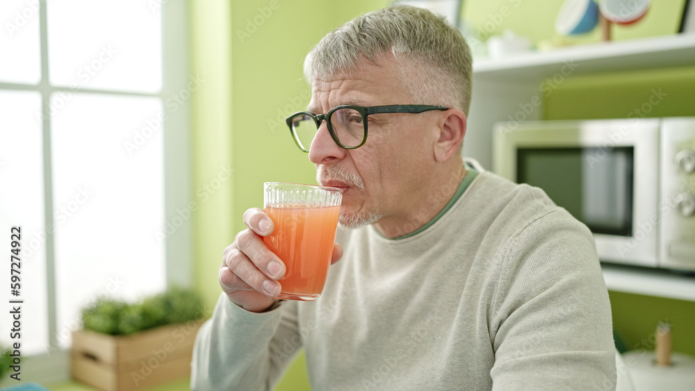 Middle age grey-haired man drinking glass of juice sitting on table at home Middle age grey-haired man drinking glass of juice sitting on table at home