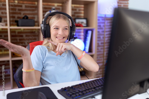 Young caucasian woman playing video games wearing headphones amazed and smiling to the camera while presenting with hand and pointing with finger.