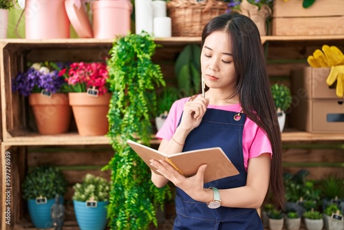 Young chinese woman florist writing on notebook with doubt expression at flower shop
