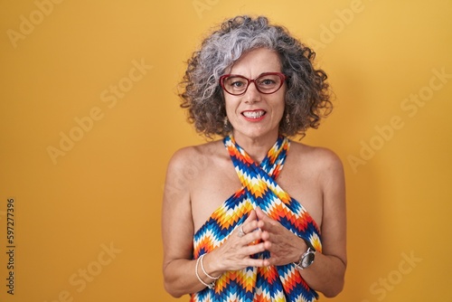 Middle age woman with grey hair standing over yellow background hands together and fingers crossed smiling relaxed and cheerful. success and optimistic