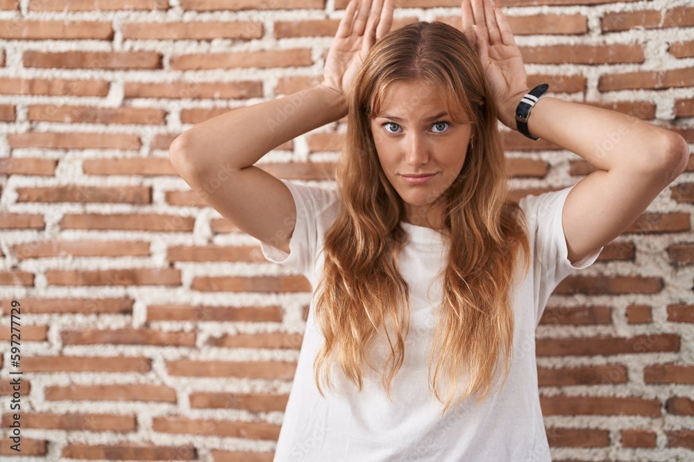 Young caucasian woman standing over bricks wall doing bunny ears gesture with hands palms looking cynical and skeptical. easter rabbit concept. Young caucasian woman standing over bricks wall doing bunny ears gesture with hands palms looking cynical and skeptical. easter rabbit concept.