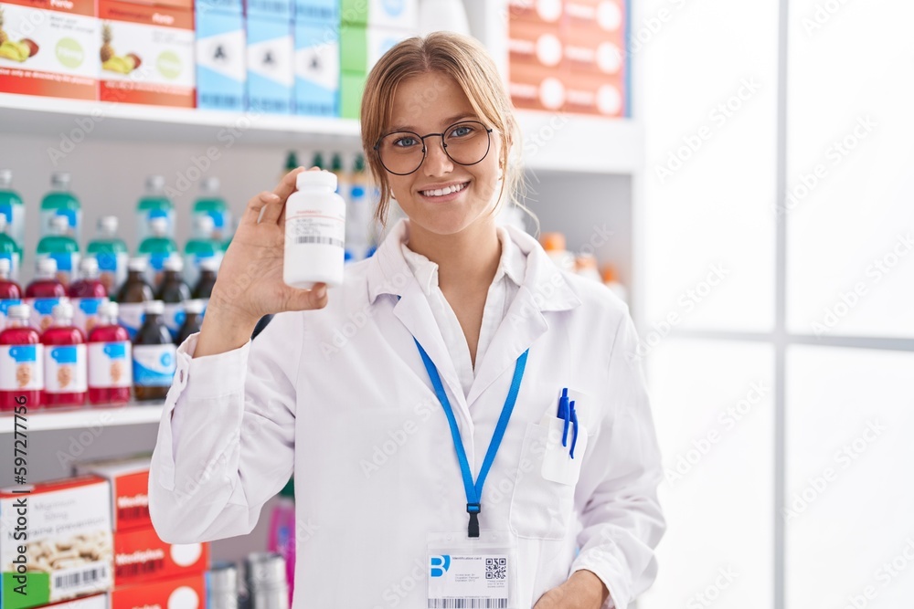 Young blonde girl pharmacist smiling confident holding pills bottle at pharmacy