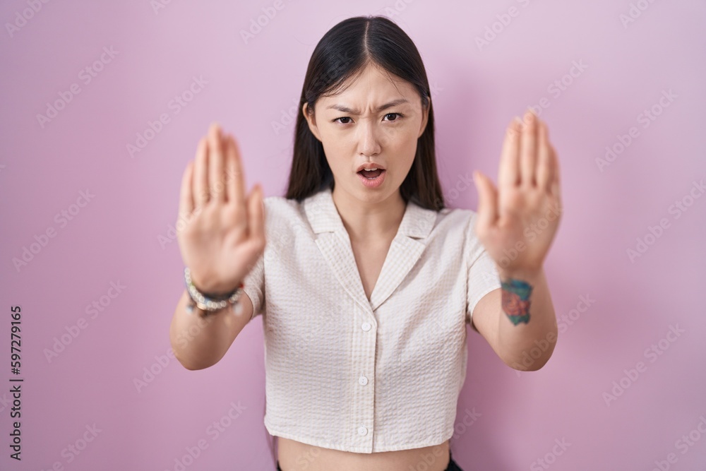 Chinese young woman standing over pink background doing stop gesture with hands palms, angry and frustration expression Chinese young woman standing over pink background doing stop gesture with hands palms, angry and frustration expression