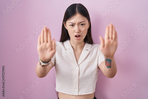 Chinese young woman standing over pink background doing stop gesture with hands palms  angry and frustration expression