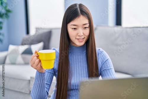 Chinese woman using laptop and drinking coffee sitting on floor at home