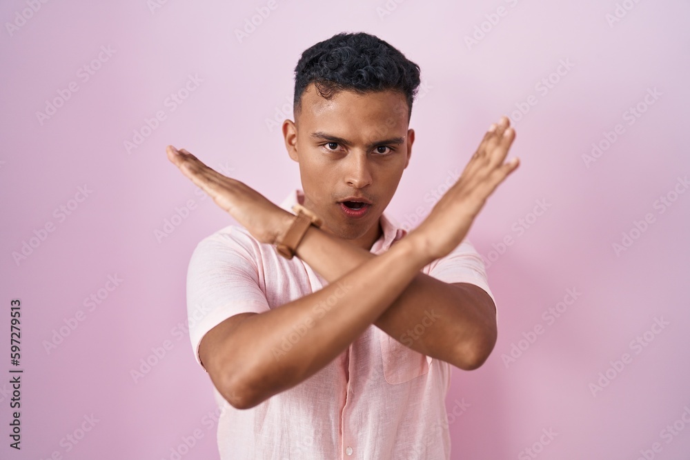 Young hispanic man standing over pink background rejection expression crossing arms doing negative sign, angry face Young hispanic man standing over pink background rejection expression crossing arms doing negative sign, angry face