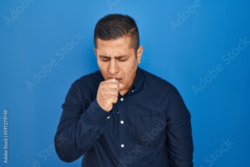Hispanic young man standing over blue background feeling unwell and coughing as symptom for cold or bronchitis. health care concept.