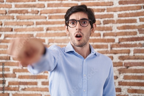 Young hispanic man standing over brick wall background pointing with finger surprised ahead, open mouth amazed expression, something on the front