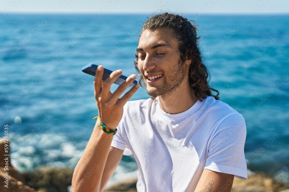 Young hispanic man talking on smartphone sitting on rock at seaside