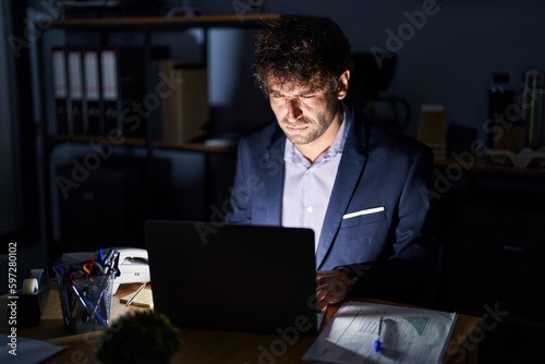 Photo Hispanic young man working at the office at night skeptic and nervous, frowning upset because of problem