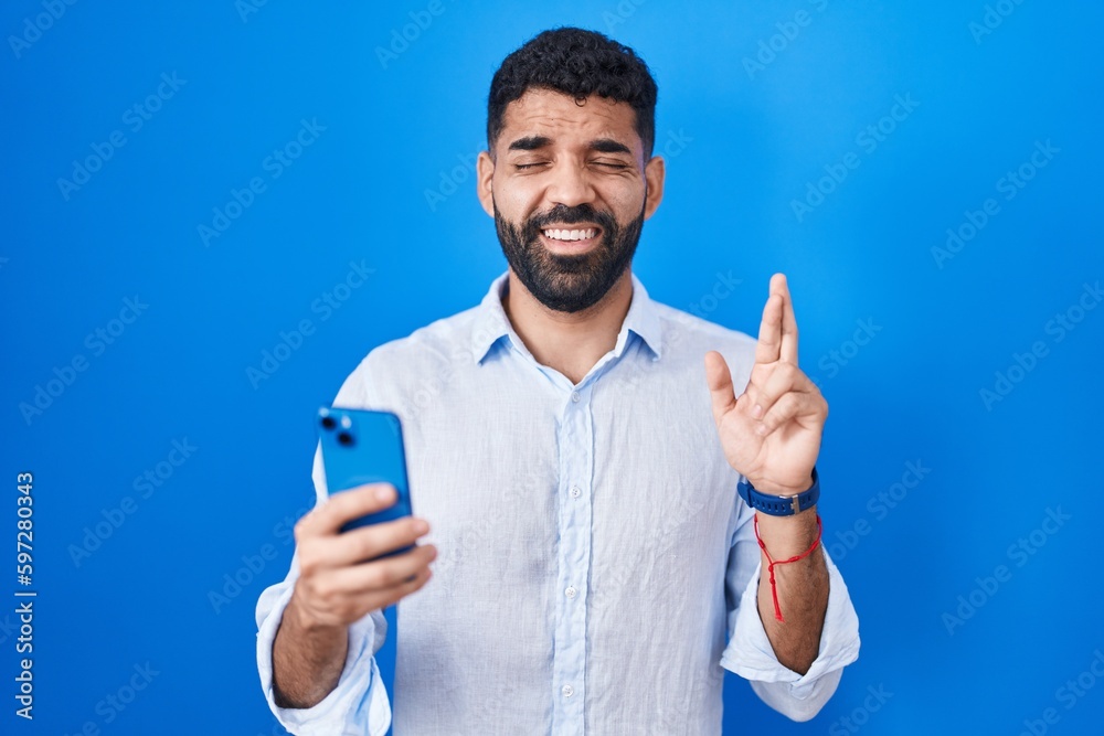 Hispanic man with beard using smartphone typing message gesturing finger crossed smiling with hope and eyes closed. luck and superstitious concept. Hispanic man with beard using smartphone typing message gesturing finger crossed smiling with hope and eyes closed. luck and superstitious concept.