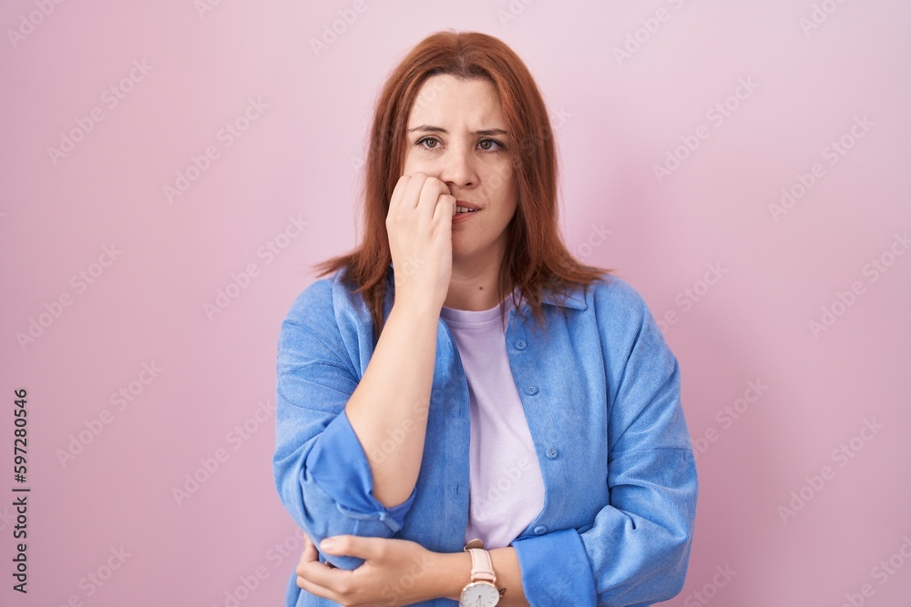 Young hispanic woman with red hair standing over pink background looking stressed and nervous with hands on mouth biting nails. anxiety problem. Young hispanic woman with red hair standing over pink background looking stressed and nervous with hands on mouth biting nails. anxiety problem.