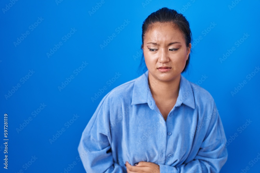Asian young woman standing over blue background with hand on stomach because indigestion, painful illness feeling unwell. ache concept. Asian young woman standing over blue background with hand on stomach because indigestion, painful illness feeling unwell. ache concept.