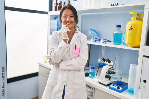 Young chinese woman wearing scientist uniform standing with arms crossed gesture at laboratory