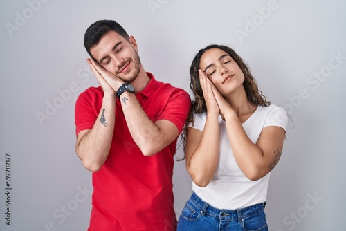 Young hispanic couple standing over isolated background sleeping tired dreaming and posing with hands together while smiling with closed eyes.