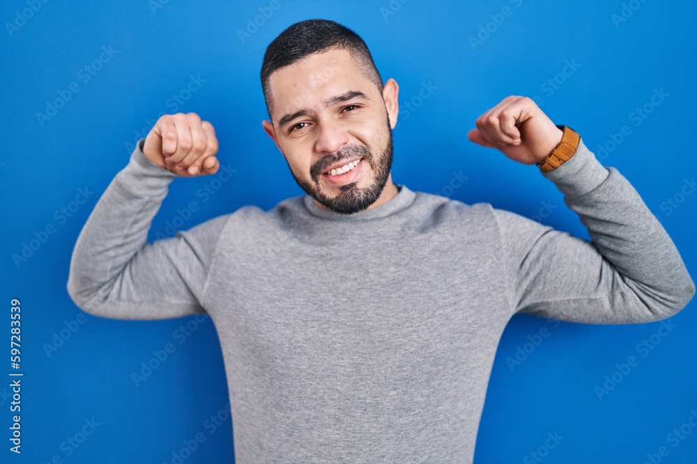 Hispanic man standing over blue background showing arms muscles smiling proud. fitness concept. Hispanic man standing over blue background showing arms muscles smiling proud. fitness concept.