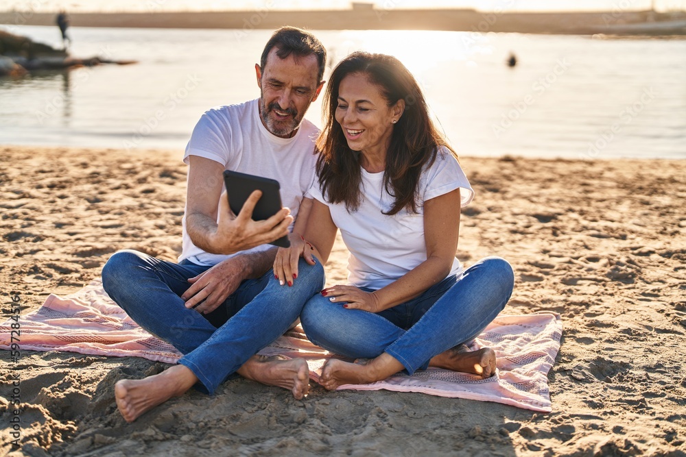 Middle age man and woman couple using touchpad sitting on sand at seaside Middle age man and woman couple using touchpad sitting on sand at seaside