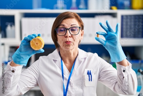 Middle age hispanic woman working at scientist laboratory making vitamin making fish face with mouth and squinting eyes  crazy and comical.