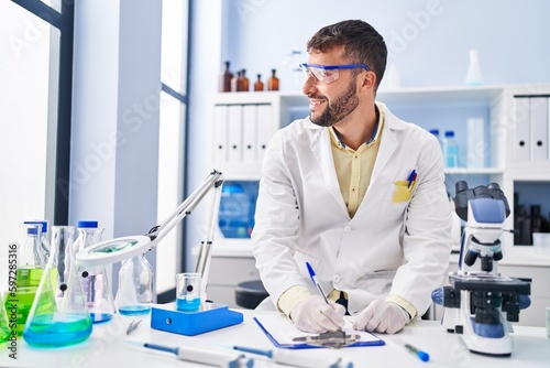 Young hispanic man wearing scientist uniform writing on clipboard at laboratory