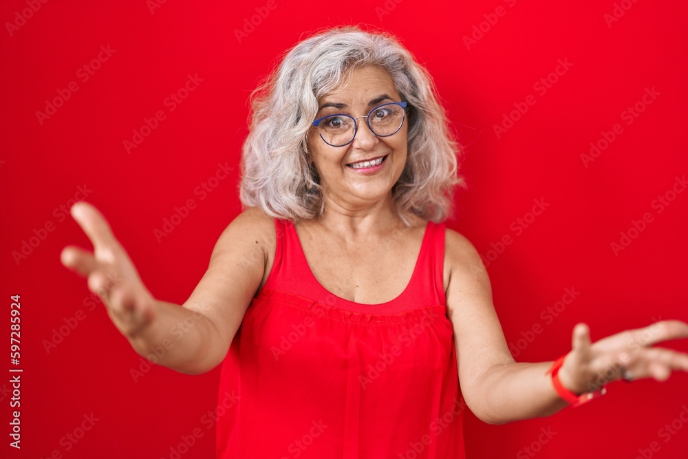 Middle age woman with grey hair standing over red background smiling cheerful offering hands giving assistance and acceptance. Middle age woman with grey hair standing over red background smiling cheerful offering hands giving assistance and acceptance.