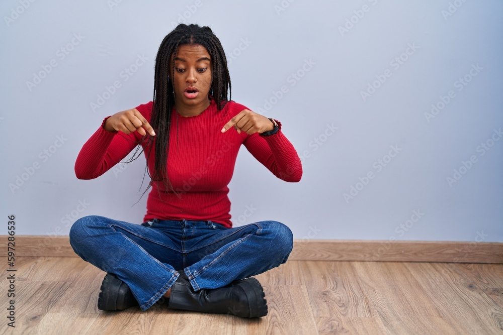 Young african american with braids sitting on the floor at home pointing down with fingers showing advertisement, surprised face and open mouth Young african american with braids sitting on the floor at home pointing down with fingers showing advertisement, surprised face and open mouth