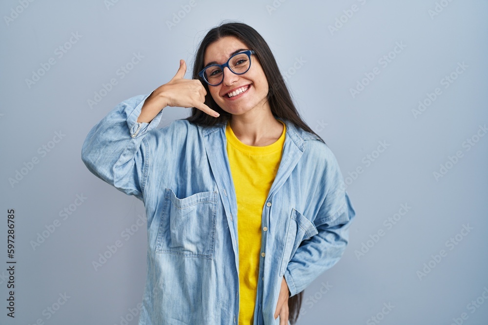 Young hispanic woman standing over blue background smiling doing phone gesture with hand and fingers like talking on the telephone. communicating concepts. Young hispanic woman standing over blue background smiling doing phone gesture with hand and fingers like talking on the telephone. communicating concepts.