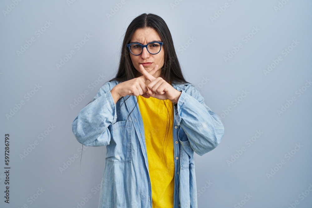 Young hispanic woman standing over blue background rejection expression crossing fingers doing negative sign Young hispanic woman standing over blue background rejection expression crossing fingers doing negative sign