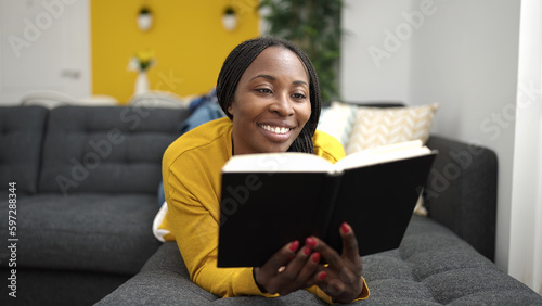 African woman reading book lying on sofa at home