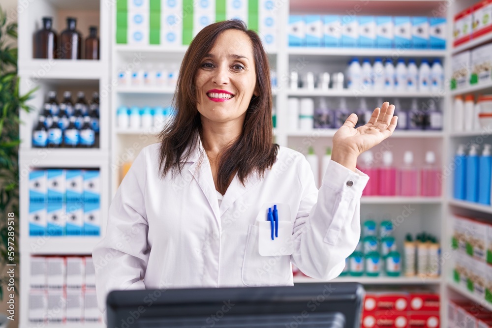 Middle age brunette woman working at pharmacy drugstore smiling cheerful presenting and pointing with palm of hand looking at the camera.