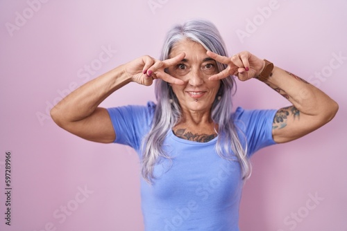 Middle age woman with tattoos standing over pink background doing peace symbol with fingers over face, smiling cheerful showing victory © Krakenimages.com