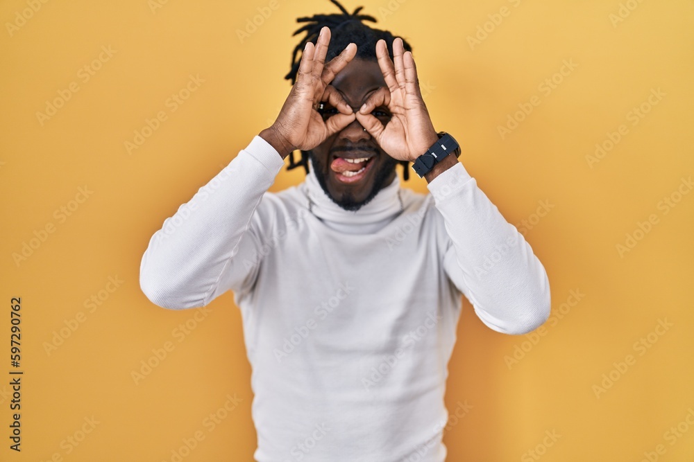 African man with dreadlocks wearing turtleneck sweater over yellow background doing ok gesture like binoculars sticking tongue out, eyes looking through fingers. crazy expression. African man with dreadlocks wearing turtleneck sweater over yellow background doing ok gesture like binoculars sticking tongue out, eyes looking through fingers. crazy expression.