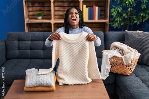 African american woman folding clean laundry angry and mad screaming frustrated and furious, shouting with anger looking up.