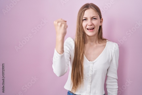 Young caucasian woman standing over pink background angry and mad raising fist frustrated and furious while shouting with anger. rage and aggressive concept.