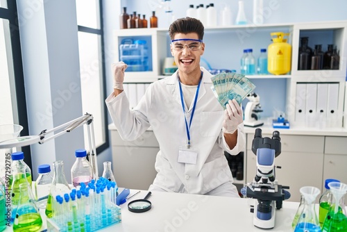 Young hispanic man working at scientist laboratory holding brazilian reals screaming proud  celebrating victory and success very excited with raised arm