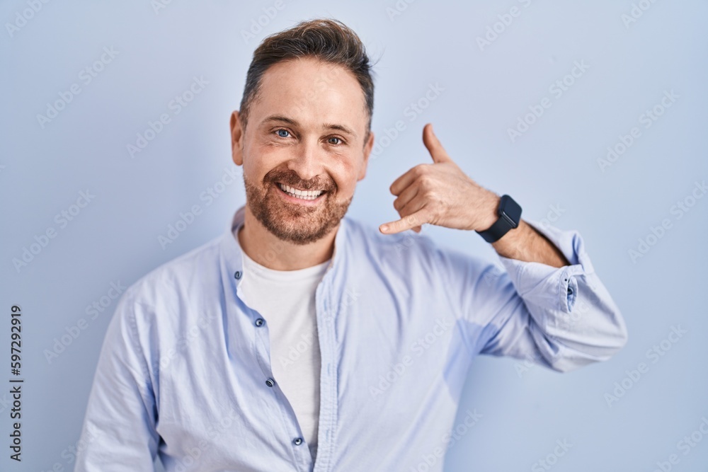 Middle age caucasian man standing over blue background smiling doing phone gesture with hand and fingers like talking on the telephone. communicating concepts. Middle age caucasian man standing over blue background smiling doing phone gesture with hand and fingers like talking on the telephone. communicating concepts.