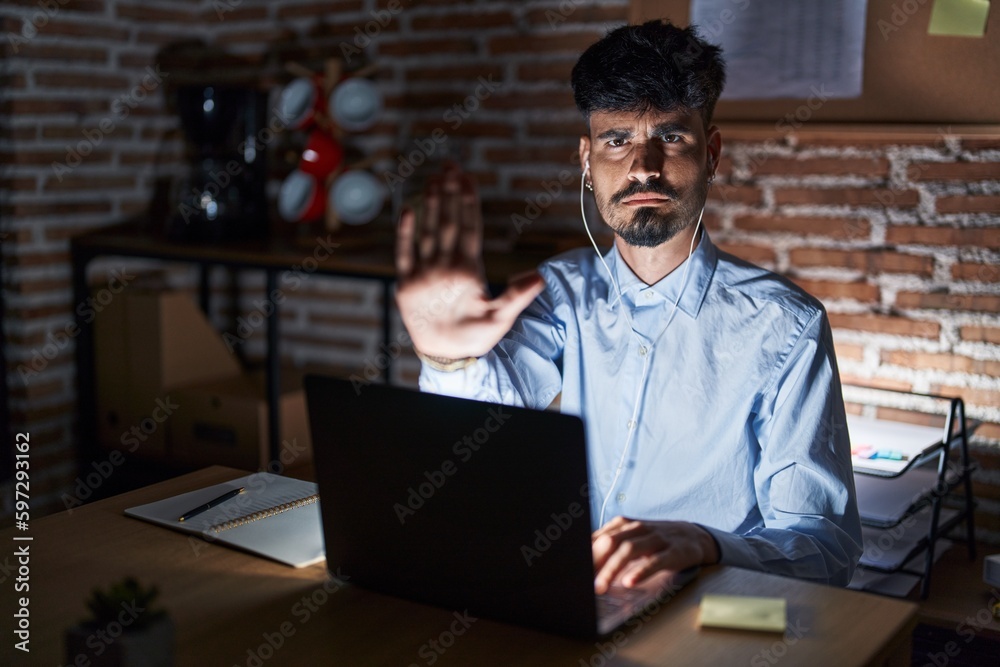 Young hispanic man with beard working at the office at night doing stop sing with palm of the hand. warning expression with negative and serious gesture on the face. Young hispanic man with beard working at the office at night doing stop sing with palm of the hand. warning expression with negative and serious gesture on the face.