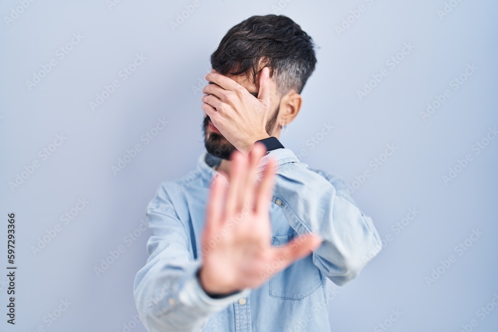 Young hispanic man with beard standing over blue background covering eyes with hands and doing stop gesture with sad and fear expression. embarrassed and negative concept. Young hispanic man with beard standing over blue background covering eyes with hands and doing stop gesture with sad and fear expression. embarrassed and negative concept.