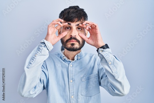 Young hispanic man with beard standing over blue background trying to open eyes with fingers, sleepy and tired for morning fatigue photo