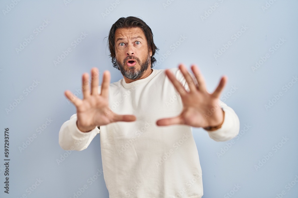 Handsome middle age man wearing casual sweater over blue background afraid and terrified with fear expression stop gesture with hands, shouting in shock. panic concept. Handsome middle age man wearing casual sweater over blue background afraid and terrified with fear expression stop gesture with hands, shouting in shock. panic concept.