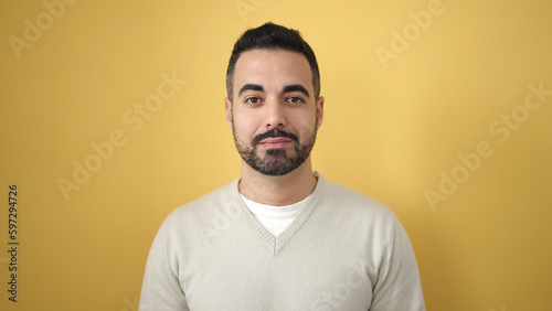 Young hispanic man standing with serious expression over isolated yellow background